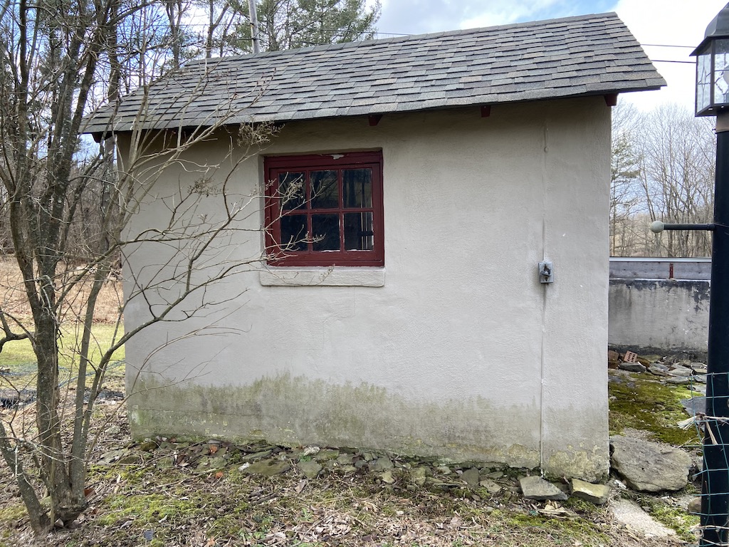 side of shed with small maroon framed window, tan shed with a lot of wear and tear on bottom near grass