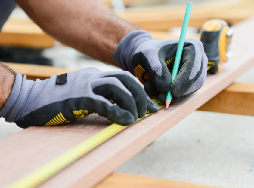 Carpenter's hands in gloves measuring wood with green pencil and tape measure