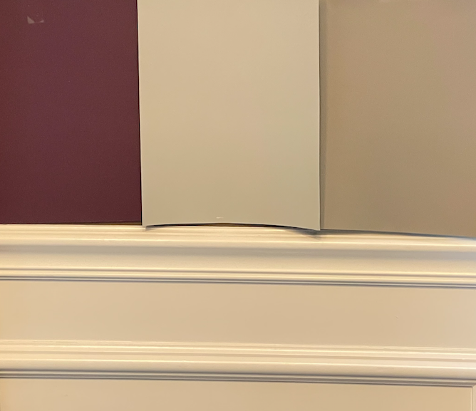 Large paint color samples in plum and beige are displayed on a brown wall with checkered curtains