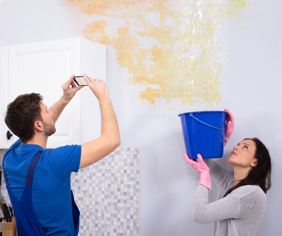 two individuals holding up bucket to collect water with water damage wall behind them